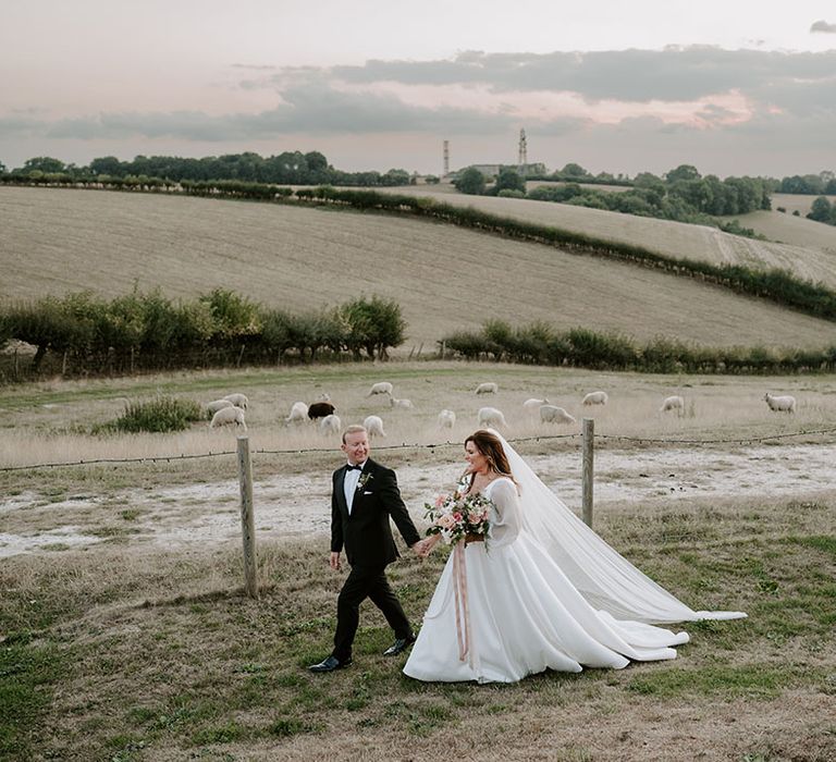 Bride & groom walk across fields as bride holds pastel bouquet tied with pink ribbon and wears sheer full sleeve Princess wedding dress