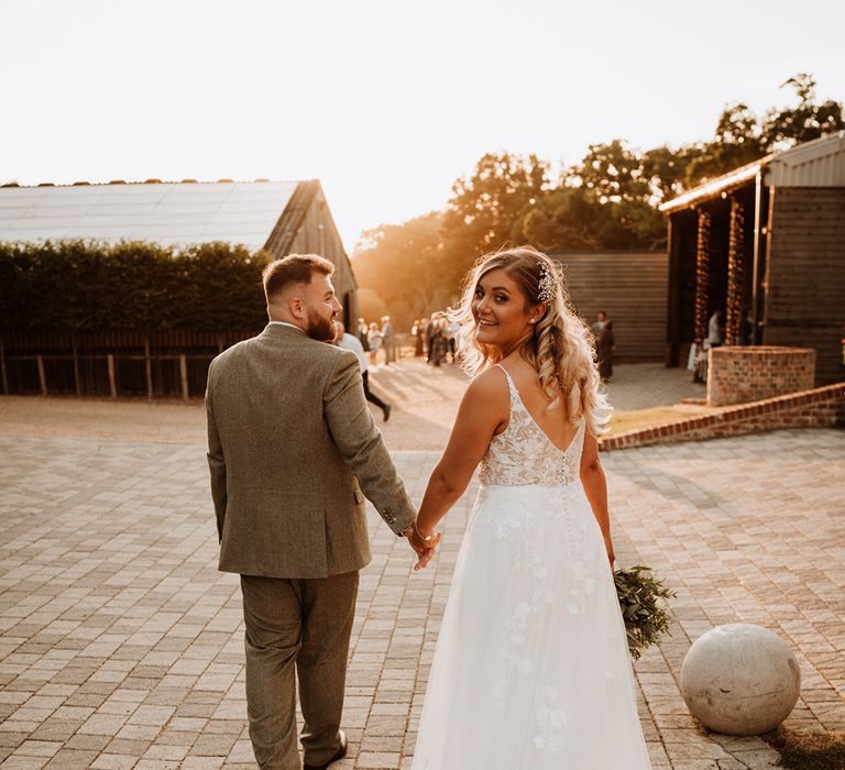 The groom in a grey suit walks holding hands with the bride who looks back at the camera wearing a sparkly hairpiece 