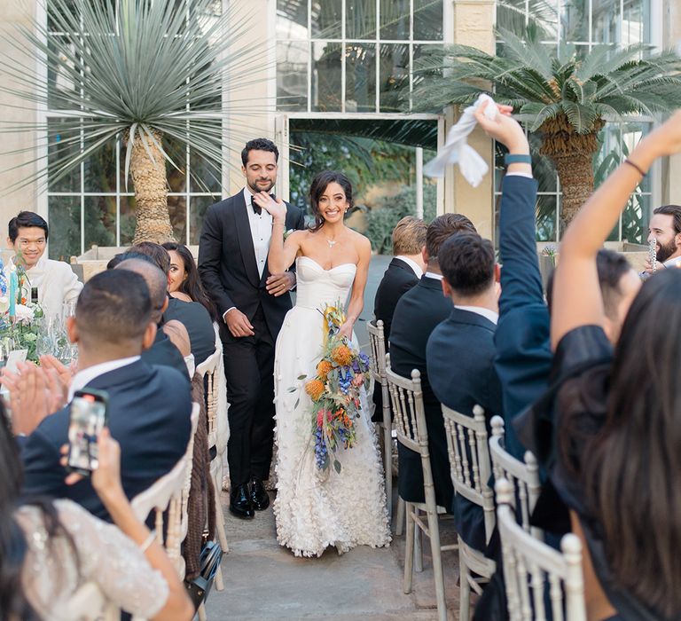 Bride holds colourful floral bouquet and walks with her groom in black-tie at Syon Park Conservatory 