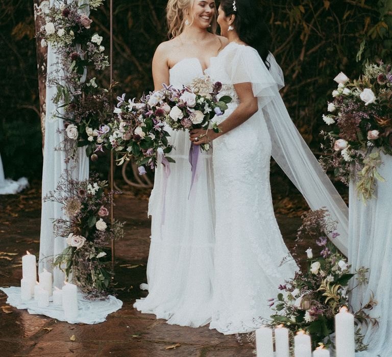 Bride in off the shoulder long sleeve wedding dress embracing bride in strapless lace wedding dress, both holding mixed neutral floral bouquets with white carnations, purple tulips, roses and foliage 