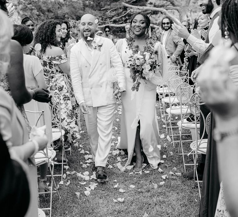 Bride and Groom walk though confetti in a cream suit and sheer-sleeve wedding dress with guests smiling and clapping