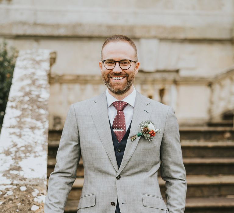 Groom in grey suit jacket with navy waistcoat with winter berries and glasses 