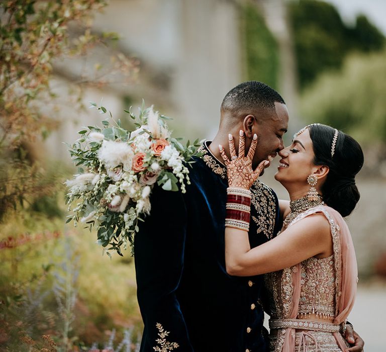 Panjabi Bride and Jamaican Groom pose at multicultural wedding. The bride wears a golden bridal Lehnga and carries a champagne bridal bouquet and the groom wears a gold embellished Indian wedding outfit