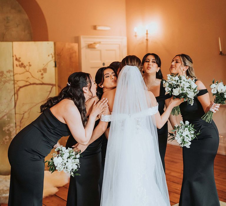 The bride receives hugs and kisses from her bridesmaids in black one shoulder dresses 