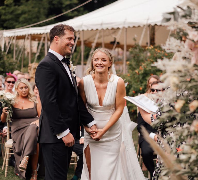 The bride laughs wearing a deep plunging wedding dress holding hands with the groom in black tuxedo for the wedding 