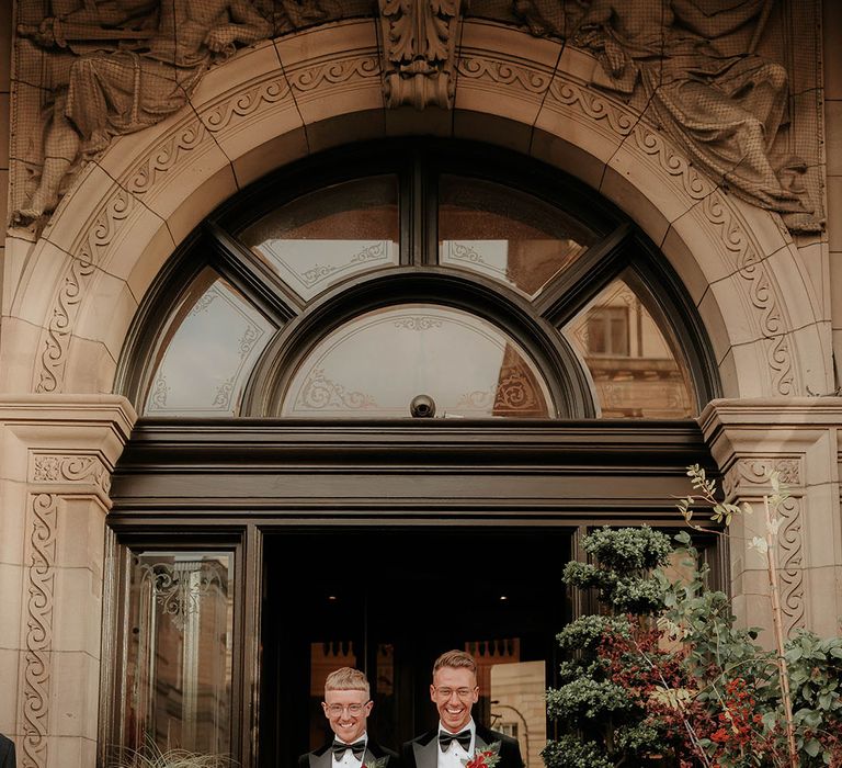 Grooms in matching velvet black tuxedos with white shirts and black bowties walking out of The Balmoral Edinburgh hand in hand with foliage arrangements in large golden and white ceramic vases on either side of them