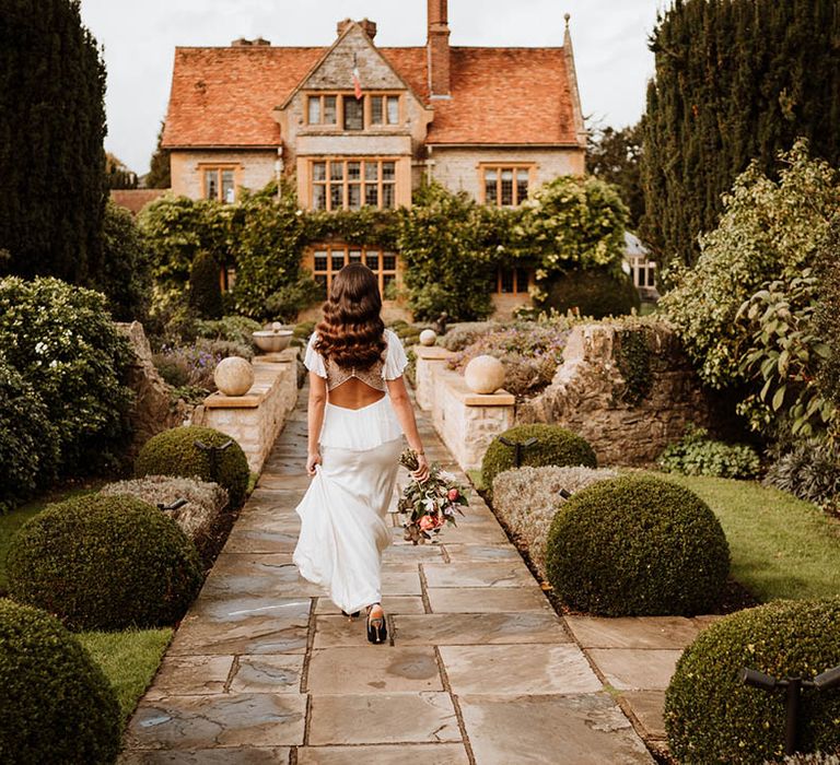 Bride in satin open back wedding dress with button details and butterfly sleeves, ruffled and layer on the back walking toward glasshouse wedding venue holding bouquet of flowers with white and pink carnations, garden roses, red strawflower, eucalyptus, baby’s-breath and various dried flowers