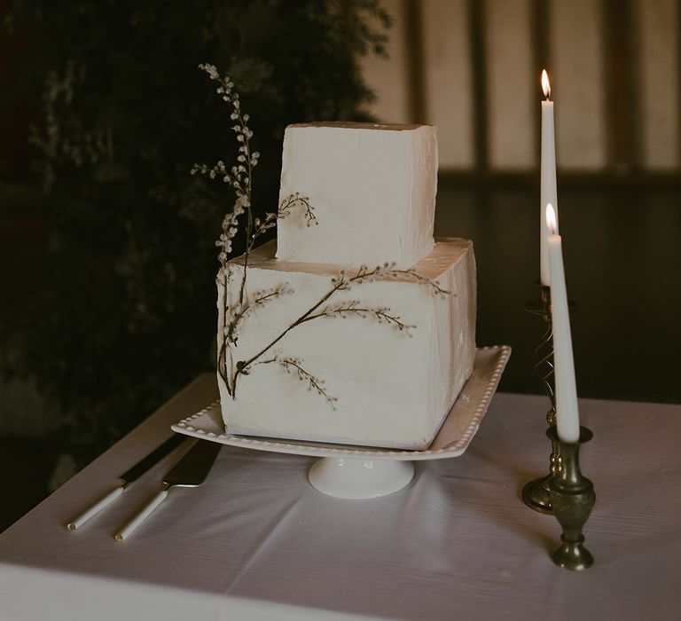 Modern two tier square wedding cake with white frosting and flower decor on white cake stand next to white taper candles 