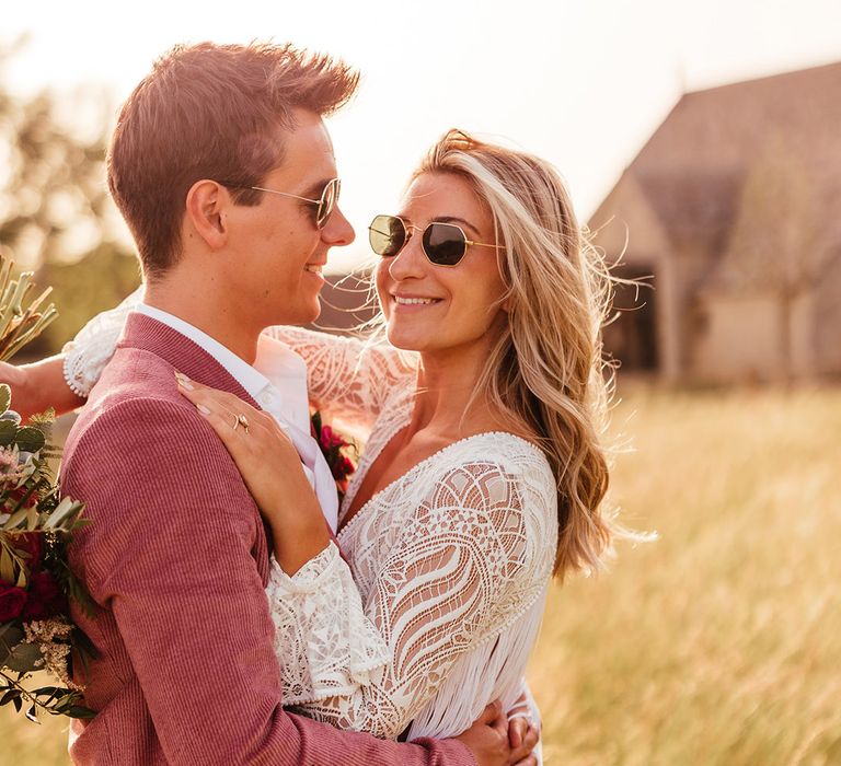 Bride in sunglasses wearing v neck long sleeve lace boho wedding dress and groom in sunglasses wearing pink corduroy suit 