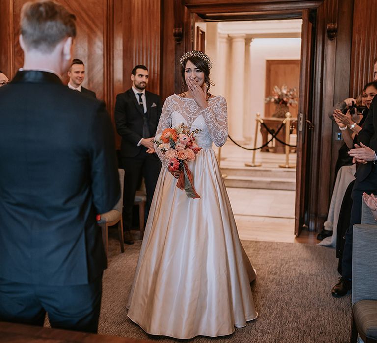 Bride walking down the aisle wearing taffeta material princess wedding dress with long lace sleeves holding rose, eucalyptus, dried flowers and Christmas berry garlands bouquet, walking towards the groom 