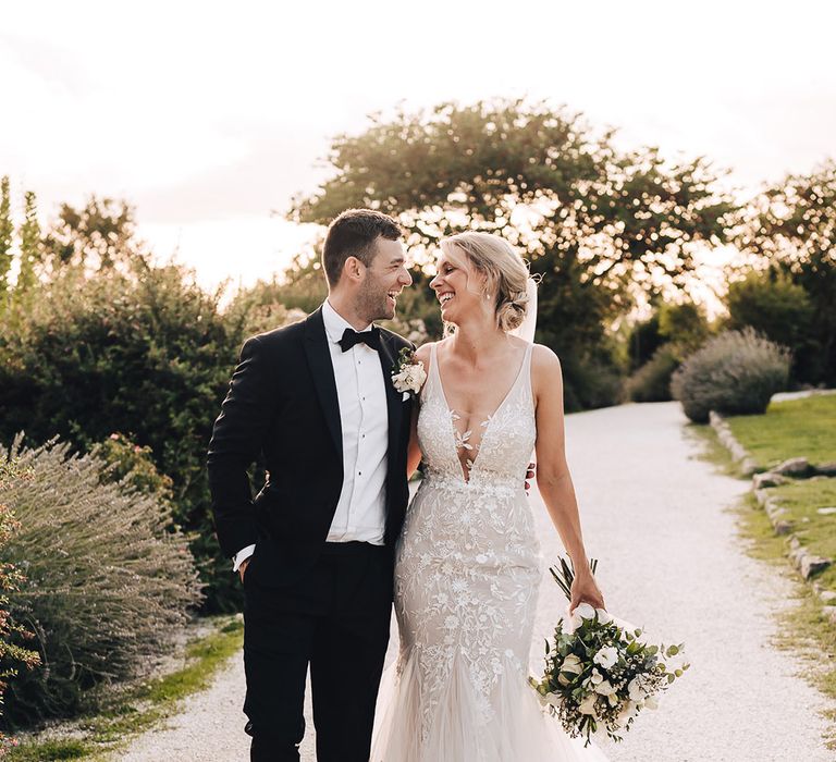 Bride in plunge lace wedding dress and trumpet styled ruffled skirt walks alongside her groom in black-tie 