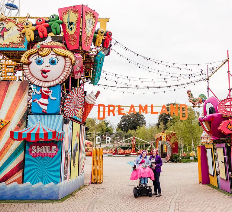 Brides standing outside Margate Dreamland theme park wedding with large colourful rides, signs and fairy lights hanging above 