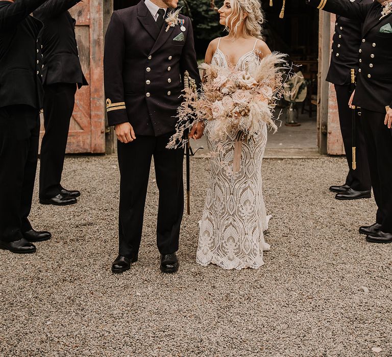 The military men make a guard of honour for the bride and groom as they look into each other's eyes with the groom in a military uniform and the bride in a boho lace gown 