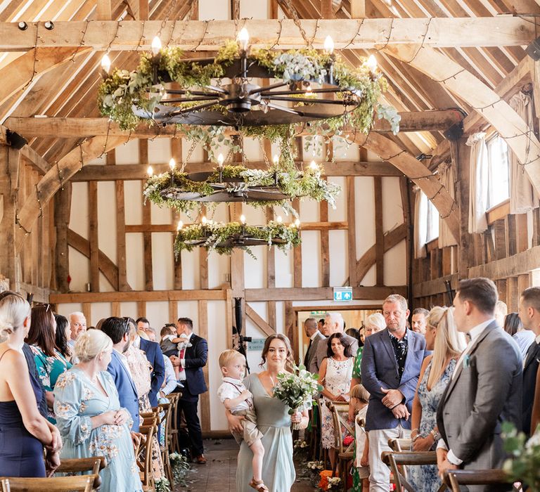 Bridesmaid in sage green bridesmaid dress walks down rustic barn aisle complete with chandeliers finished with green foliage decor 