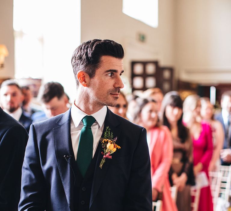 Groom in three piece navy blue suit and green tie turns his head as the bride starts walking down the aisle 