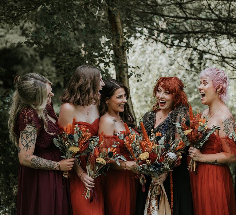 Bridesmaids in mismatched orange bridesmaid dresses and bride in v-neck embellished black dress for wedding in Devon, all holding dried flower wedding bouquets