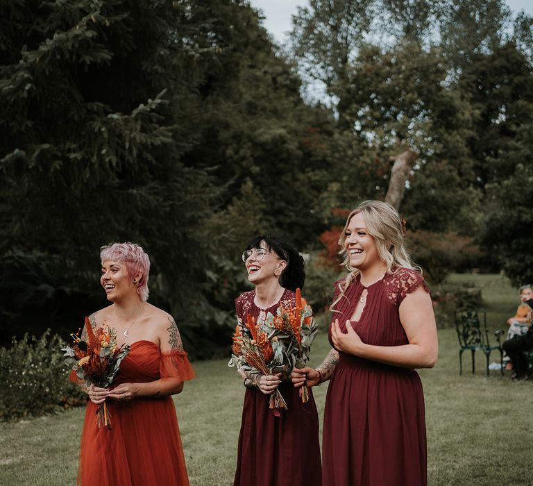 Bridesmaids in mismatched burgundy and burnt orange bridesmaid dresses holding burnt orange dried flower bouquets