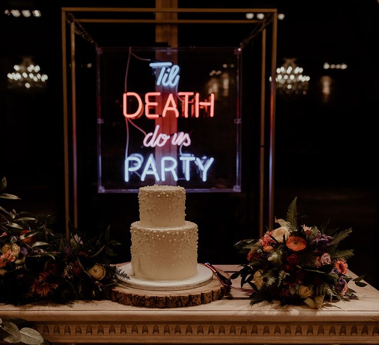 Two tier white wedding cake with pearl decoration on a tree trunk slice cake stand with neon wedding sign behind the cake 