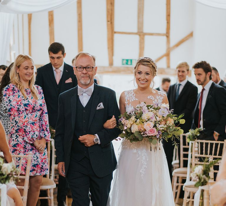 Bride in lace and tulle wedding dress holding pastel wedding bouquet being walked down the aisle by the father of the bride in a navy suit 