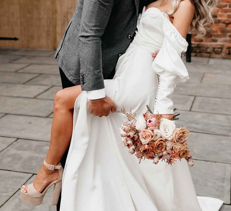 Groom in grey blazer, black waistcoat and navy polkadot tie holding bride in off the shoulder long sleeve wedding dress with slight cowl neck and front leg slit