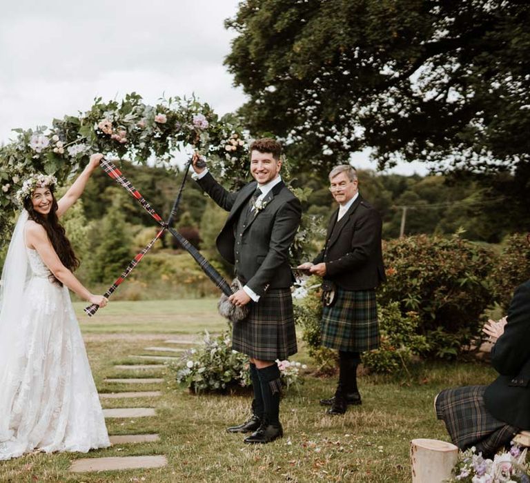 Groom in dark grey blazer and waistcoat, dark tartan tie with white flower boutonniere, dark grey and blue tartan kilt and wedding sporran doing wedding handfasting ceremony with bride in 3d appliqué sheer overlay wedding dress and bridal flower crown at The Barn at Harburn