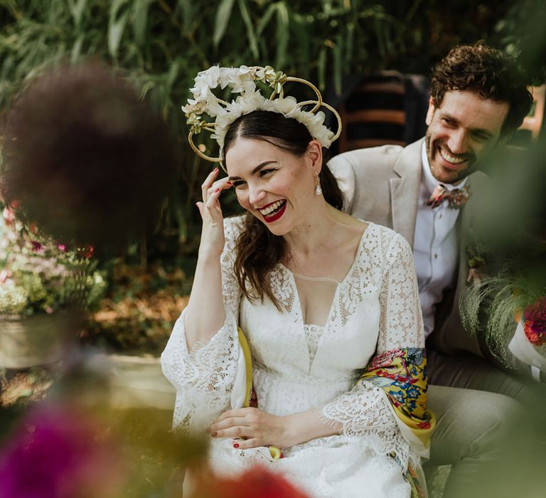 Bride wearing a lace wedding dress with a boho bridal crown and floral yellow pashmina sits at oudoor wedding with groom in greige suit