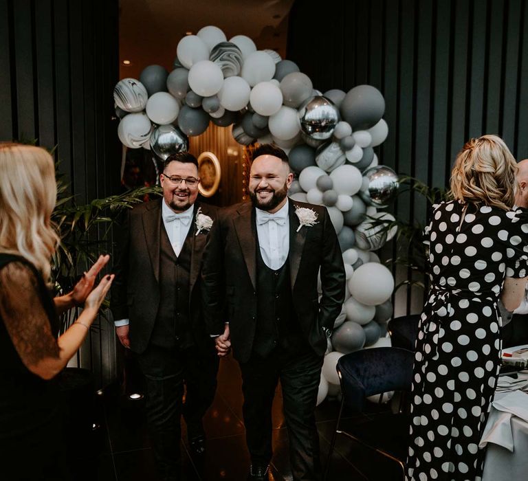 Grooms in matching classic black three piece tuxedos with white rose boutonnieres walking into the party room at The Gherkin wedding venue 