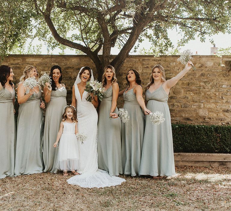 Bridesmaids in matching sage green bridesmaid dresses with the flower girl in a white dress waving their bouquets