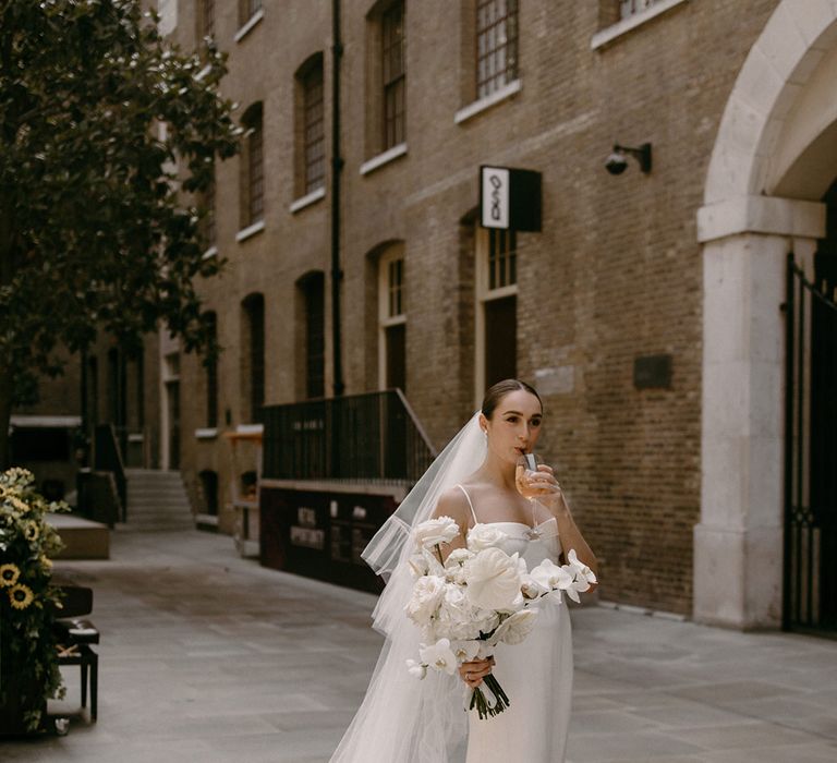 Bride in satin wedding dress with cathedral length veil with ruffle detail sipping on alcoholic drink after the ceremony 