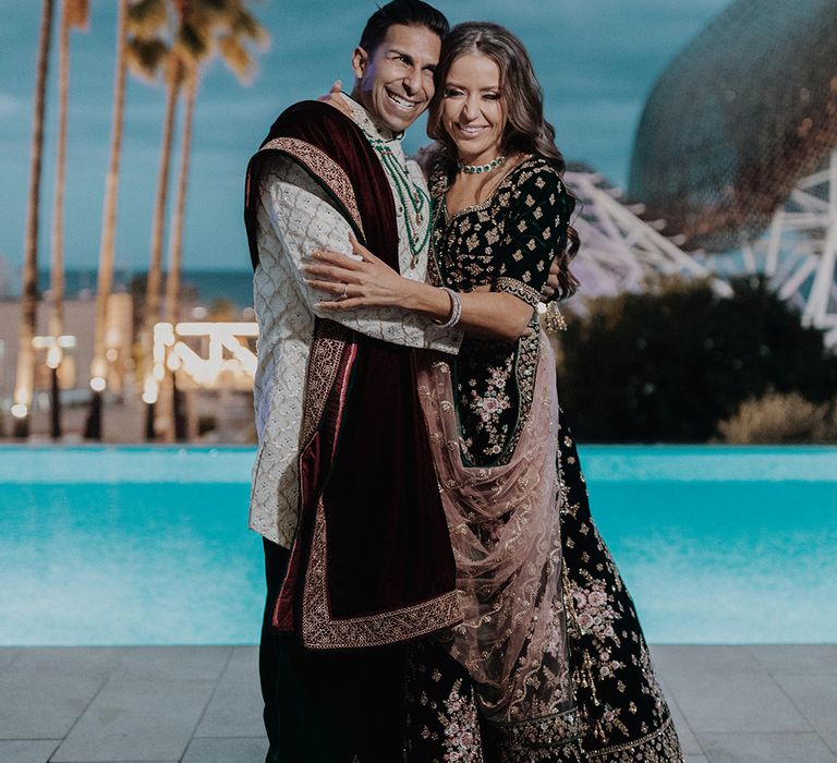 Bride and groom in black and gold traditional Indian wedding outfits posing by the pool of Spanish castle wedding venue 