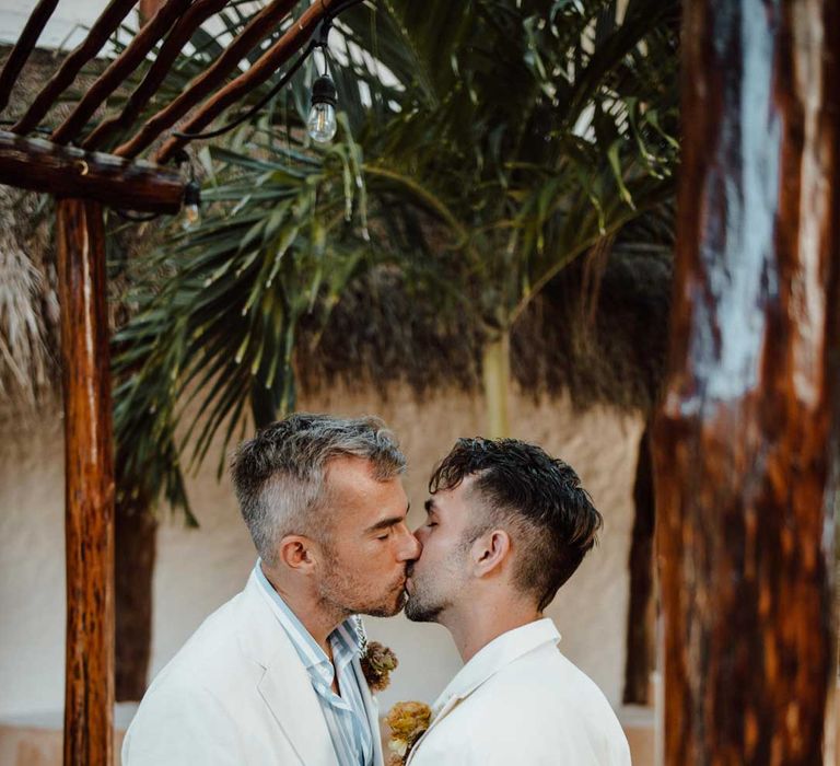 Groom in white mens wedding suit, light blue and white striped shirt, dusky orange garden rose and dried flower boutonniere, white suit trousers and gold jewellery kissing groom in white blazer with dusky orange garden rose and dried flower boutonniere, white shirt, white suit trousers and silver jewellery 