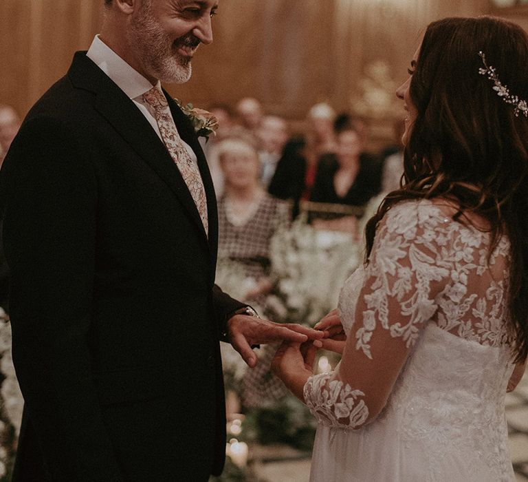 Groom in dark suit smiles at the bride as they exchange rings for the wedding ceremony with the bride in a lace wedding dress with sparkly hair accessory