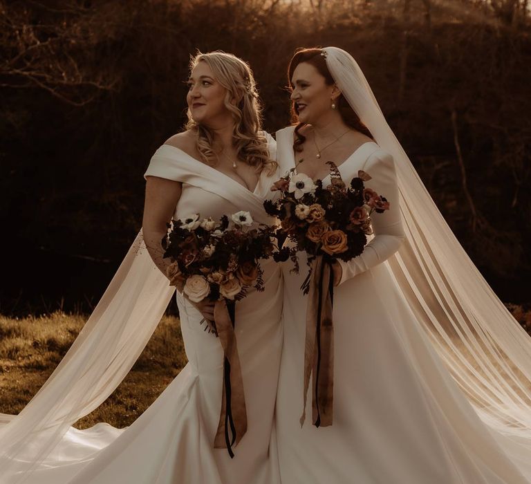 Bride in satin short sleeve deep v wedding dress and church length veil and bride in long sleeve deep v wedding dress and church length veil holding white, dusky pink and burnt red dried flower, rose and peony bridal bouquets at Hidden River Cabins golden hour photoshoot 