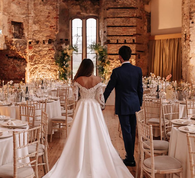 Bride in classic long sleeve wedding dress with long train walking around the wedding reception with groom in blue suit 