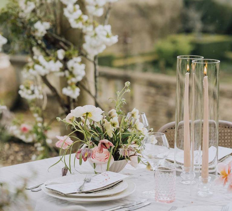 Outdoor al fresco wedding tablescape with blush pink tapered candles in glass candlestick covers on spring inspired wedding tablescape with white ruffled wedding table cloth, white peony, pink garden rose, foliage and blush florals wedding table centrepieces and simple crockery at Euridge Manor wedding