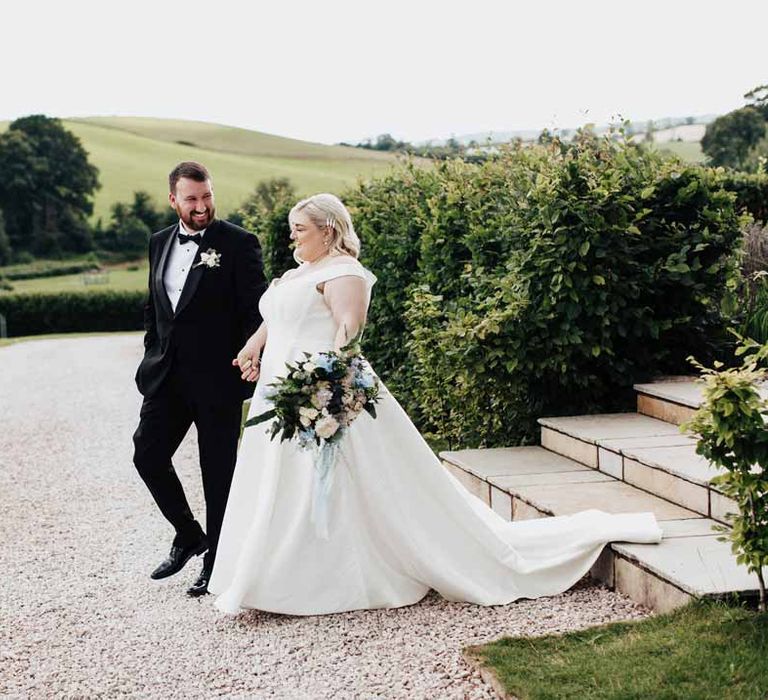 Bride in satin off the shoulder wedding dress holding white and blue eucalyptus, foliage, garden rose, gardenia, light blue delphinium and dried flower bridal bouquet tied with dusky blue ribbon holding hands with groom in classic black grooms three piece tuxedo with white rose boutonniere and black bowtie 