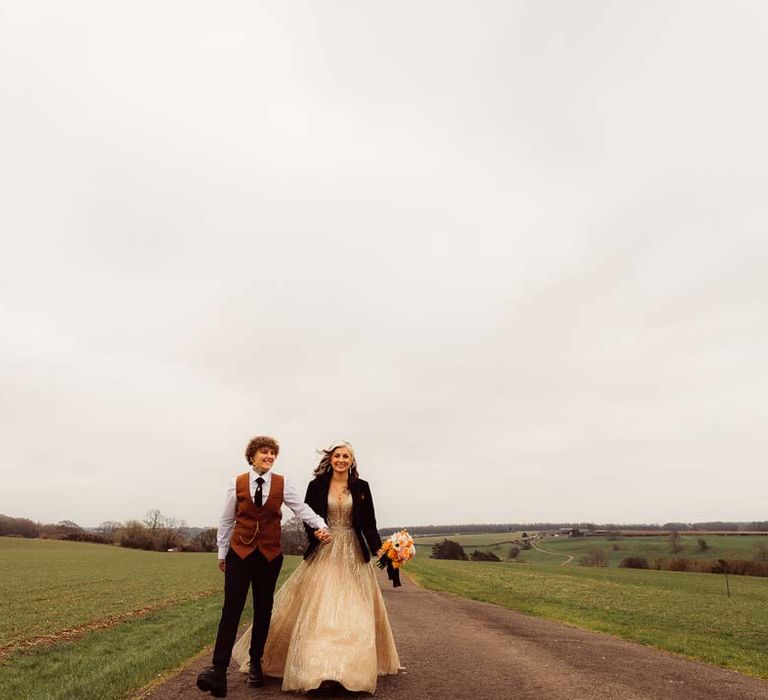 Bride in rust waistcoat and pocket square, black suit trousers, platform Dr Martens and gold pocket watch walking hand in hand with bride in sleeveless deep v-neck sparkly gold wedding dress with personalised bridal leather jacket 