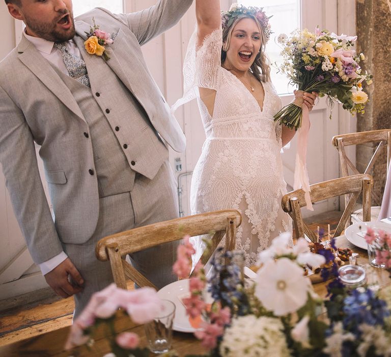 Groom in three piece light grey suit with blue and white paisley tie raising the bride's hand at their wedding breakfast with bride in lace gown 