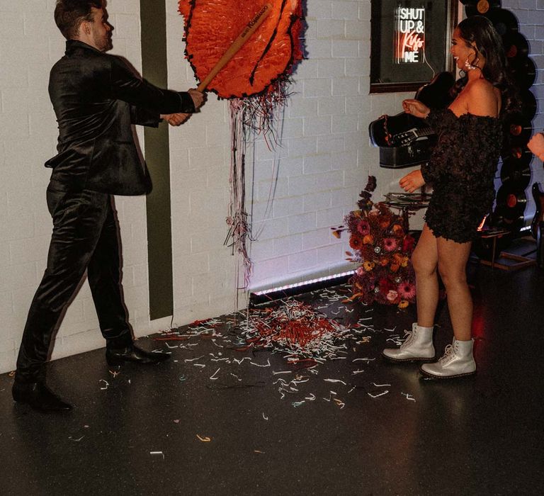 Bride in black ruffle off shoulder reception dress and red drip effect sunglasses with groom in black shirt and black drip effect sunglasses batting the Rolling Stones wedding piñata 