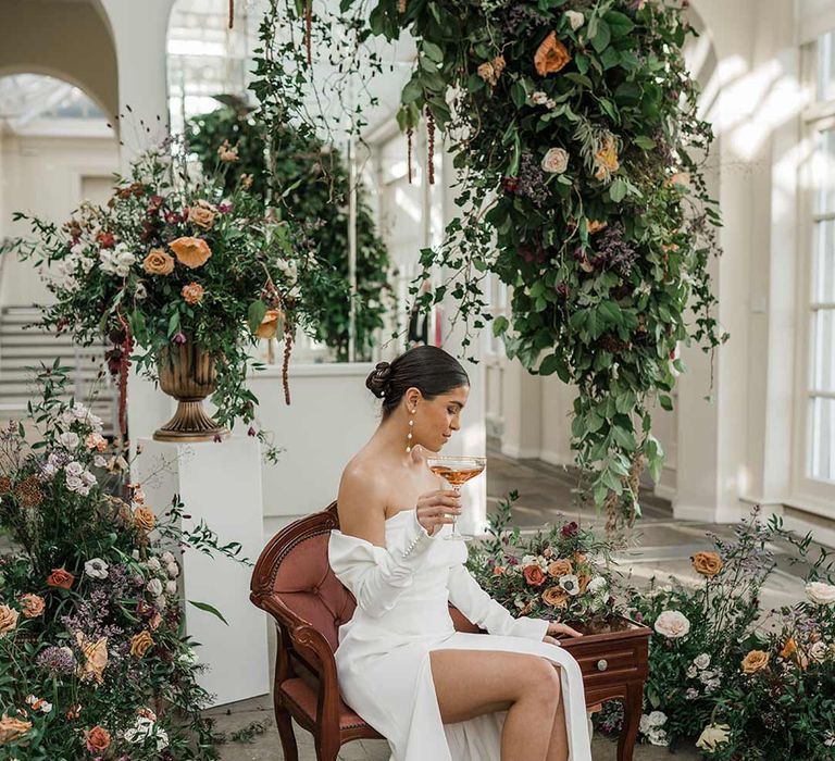 Bride in long sleeve off the shoulder wedding dress with pearl button detailing and a side slit sitting on red vintage chair at Buxted Park surrounded by suspended wildflower and foliage arrangements 