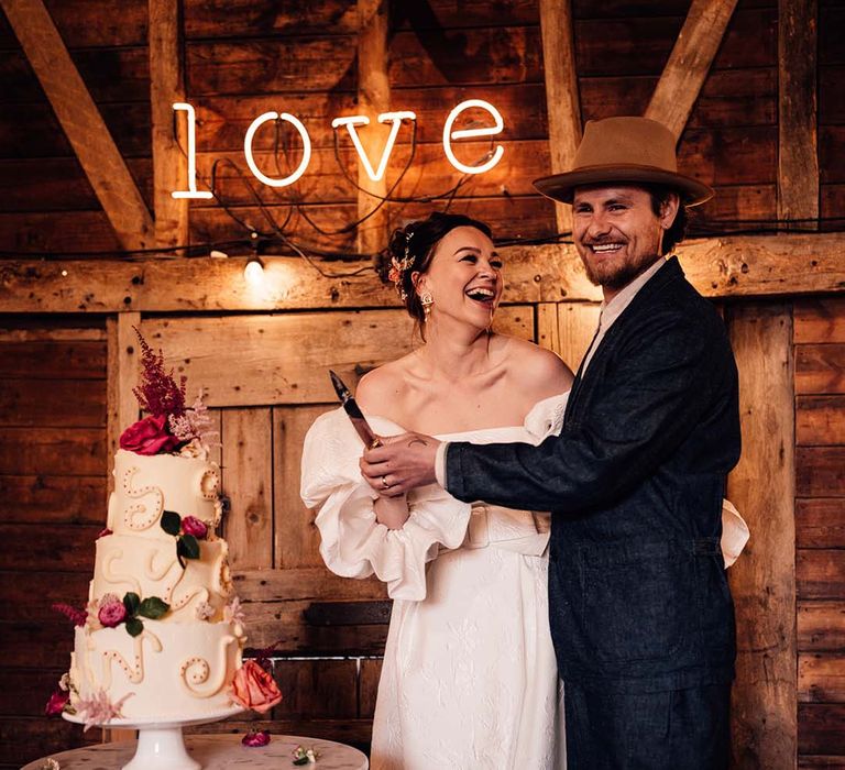 Bride and groom cut their three tier wedding cake with pink flower decorations and decorative squiggles in front of neon sign 