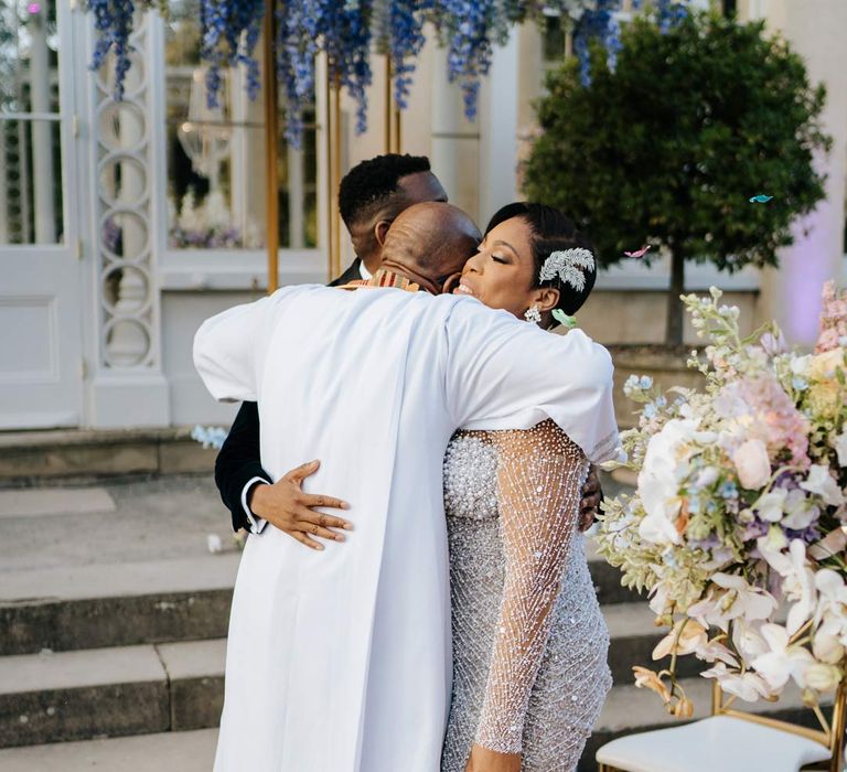 Bride in illusion v-neck sheer off shoulder long sleeve pearl embellished wedding gown being hugged by groom and officiant at Syon Park vow renewal