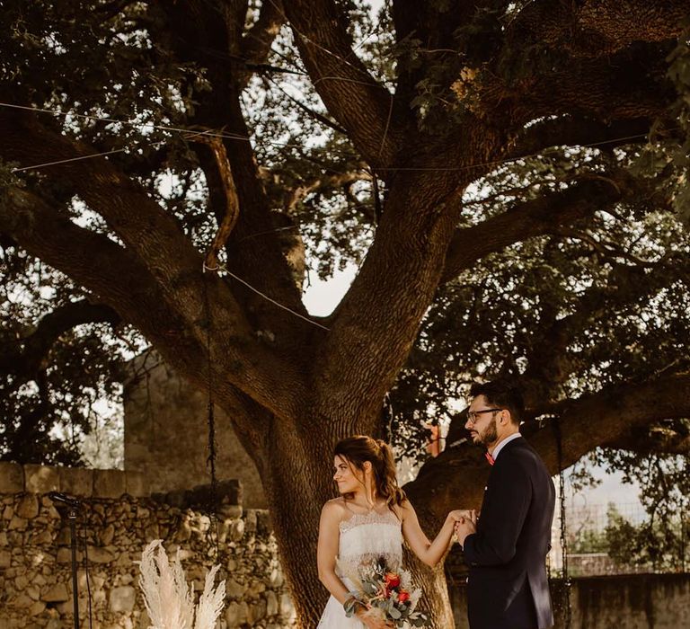 groom in grey chinos and a navy blazer holding hands with his bride in a strapless lace wedding dress at their outdoor boho wedding ceremony with pampas grass and wool rugs