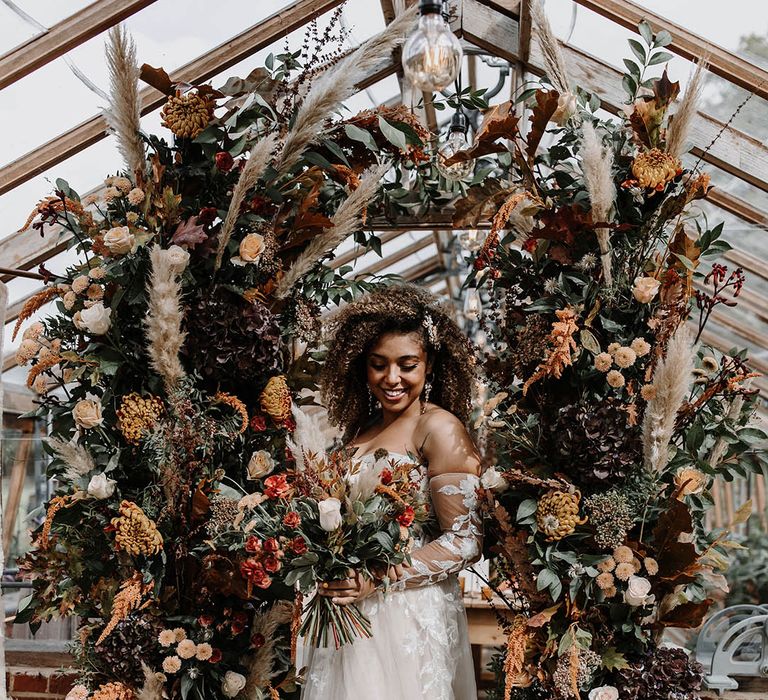 Glasshouse wedding with bride posing in front of two orange autumnal wedding flower columns 