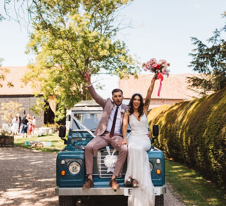 Bride and groom sit on blue Land Rover car decorated with white ribbon 