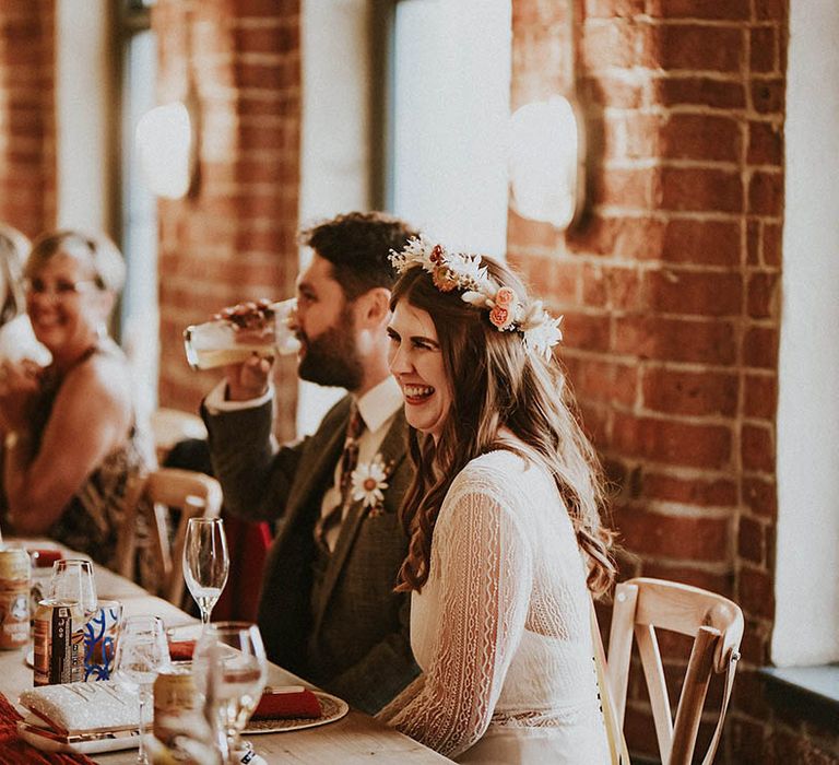 The bride wearing a flower crown sitting with the groom in a brown suit at their wedding breakfast 