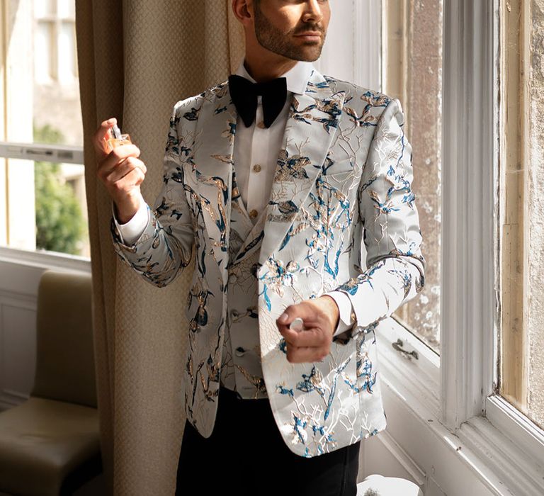 Groom in pastel blue patterned suit jacket with black bow tie putting on cologne 