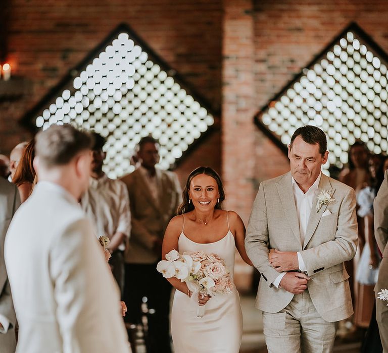 Bride in white satin wedding dress with father of the bride in grey wedding suit walk down the aisle together 