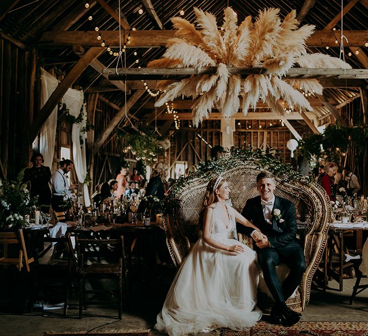 Bride and groom sitting on a peacock chair made for two under a pampas grass installation