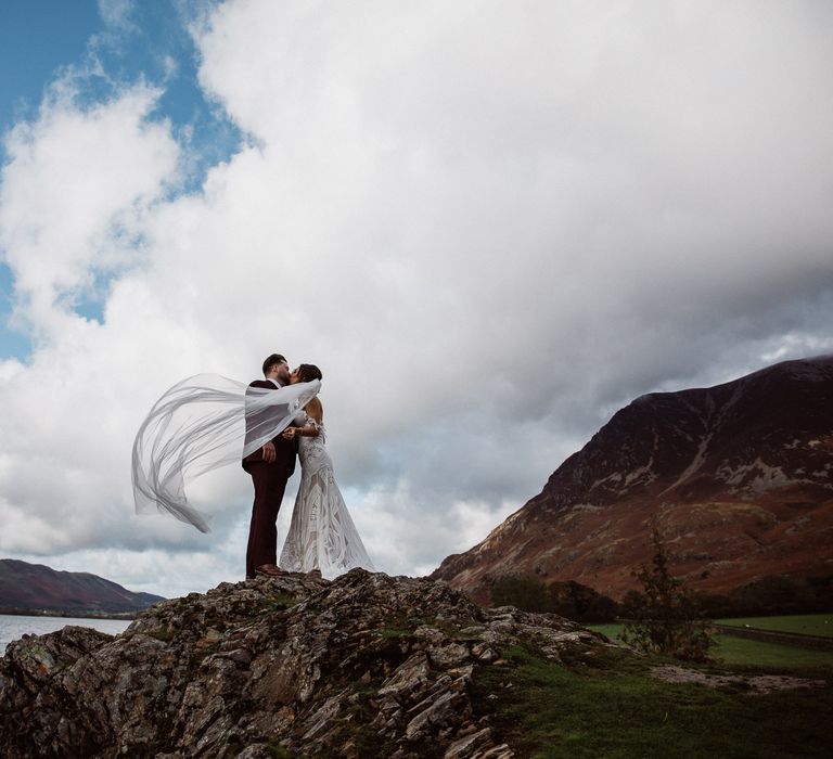 Bride with veil caught in the wind kisses Groom on a rock next to a lake